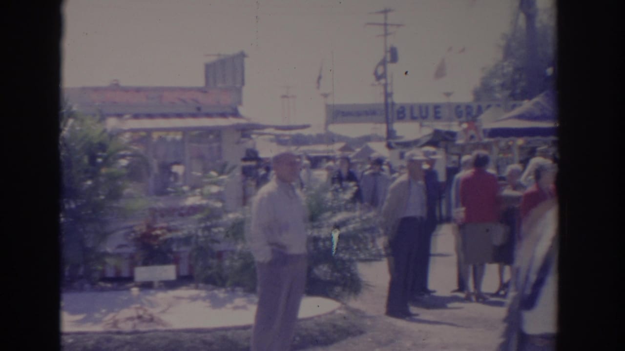 Fairgoers walking past plants and vendor booths with a partial BLUE GRASS sign behind them.