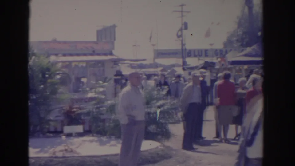 1962 Florida fairground scene with adults in period clothing walking near vendor booths, plants, flags, and a partial BLUE GRASS sign.