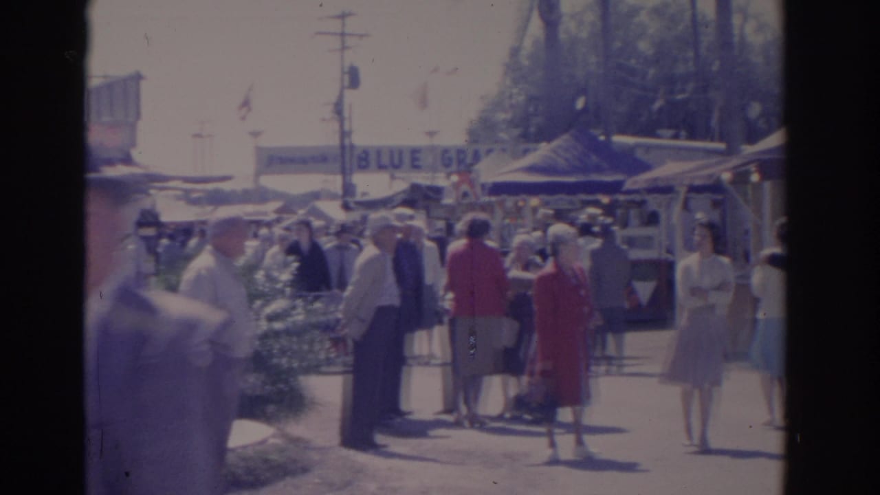 Crowd of adults standing and walking near tents, booths, and a partial BLUE GRASS sign.