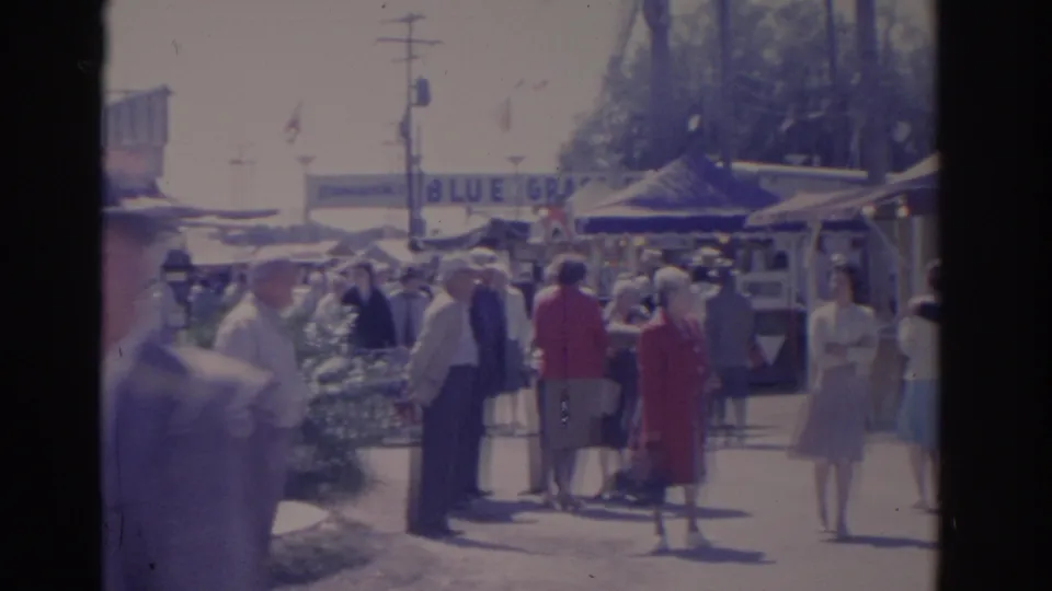 Adults in 1960s clothing gather and pass through a Florida carnival area with tents, booths, flags, and visible fair signage.