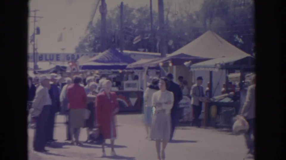Handheld 8mm view of a crowded 1962 Florida fair walkway with adults passing vendor tents and midcentury outdoor signage.