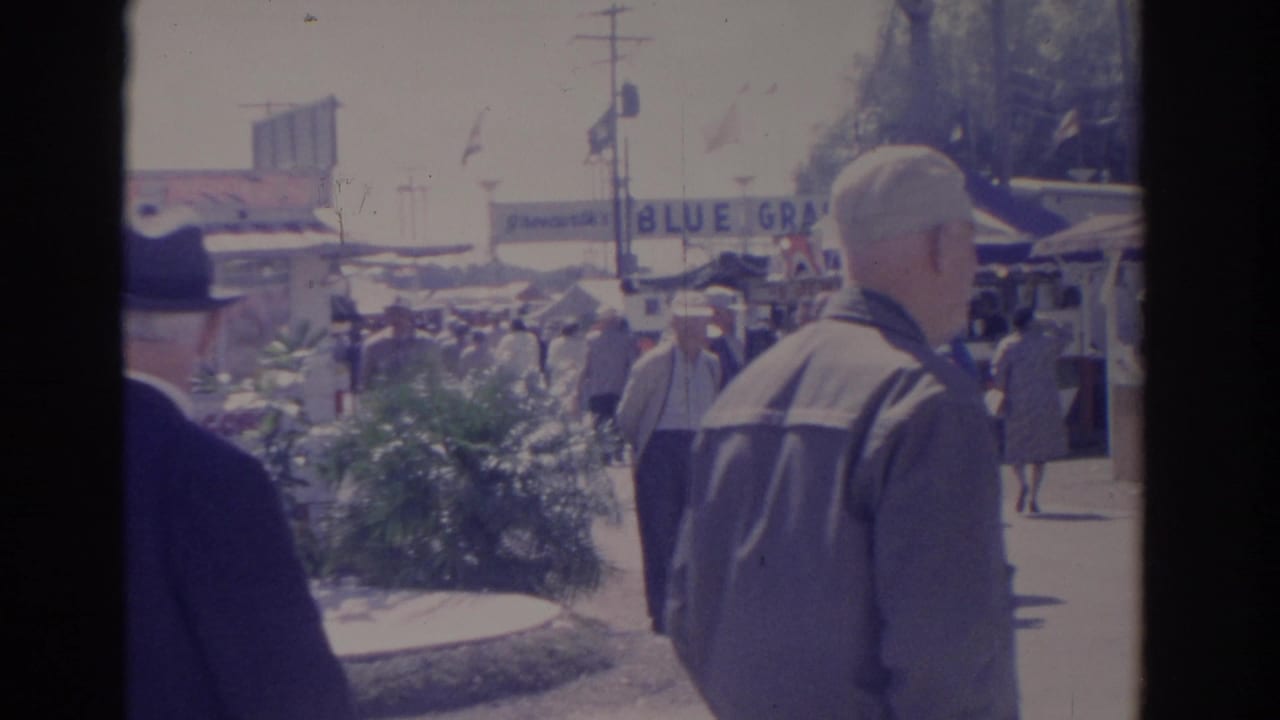 Two men seen from behind walking toward a crowded fair midway with tents and a Blue Grass sign.