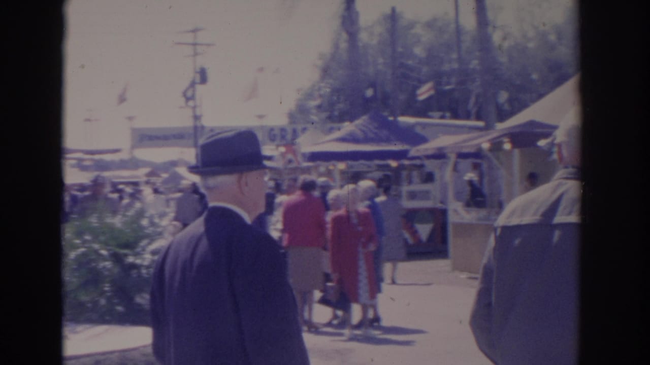 1962 Florida fair footage frame with a dark-suited man in a fedora walking past booths, tents, and other visitors.