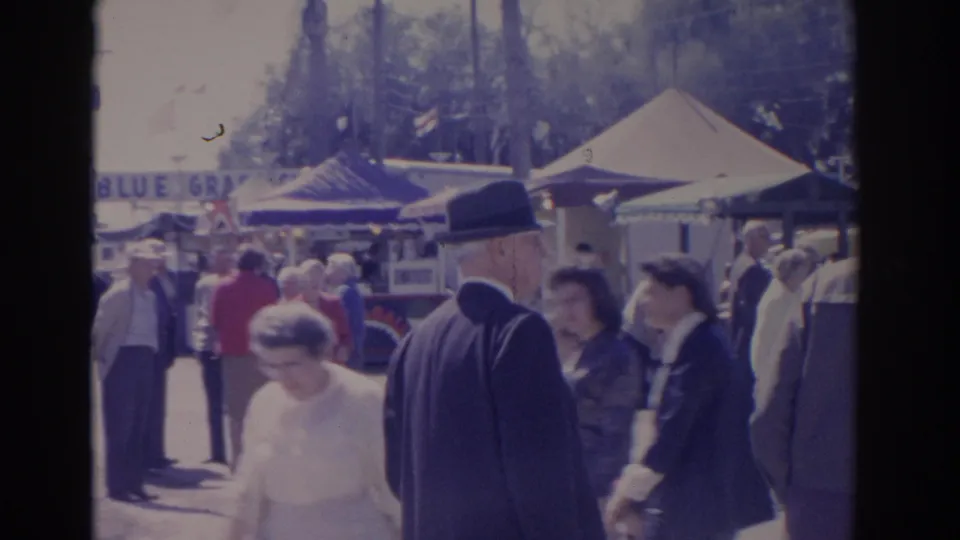 1962 Florida 8mm frame showing passing fair visitors, a suited man in a hat, tents, and a partial Blue Grass sign.