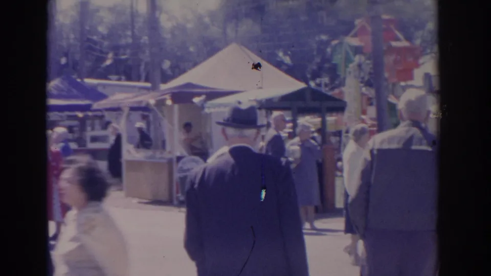 1962 Florida fair frame with a fedora-wearing man walking away near booths, canvas tents, fairgoers, and a colorful structure.