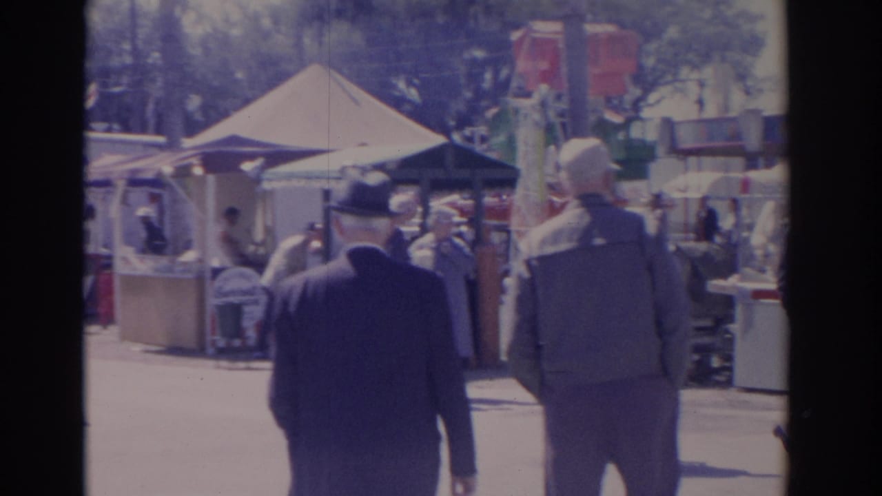 Two men walk away from the camera through a sunny carnival with tents and booths.