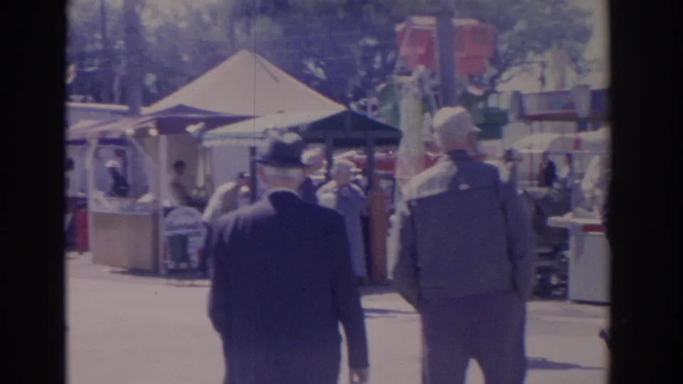 Two men in suits and hats walk through a Florida carnival in 1962, with tents, booths, and a ride visible ahead.