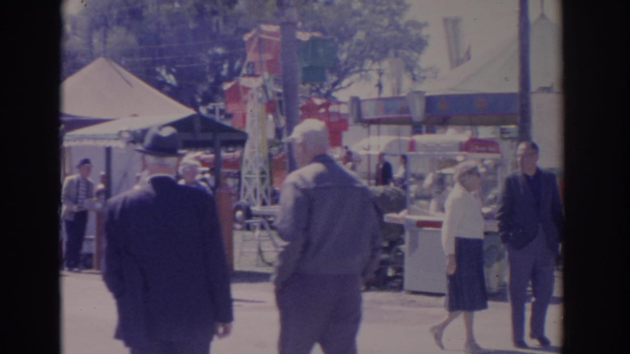 Fair visitors walk near booths and tents while two men continue through the carnival.