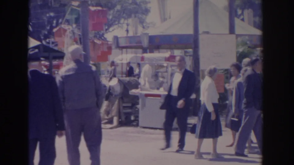 Adults in mid-century clothing walk between Florida carnival booths and tents, with a ride structure and vendor counters in the background.