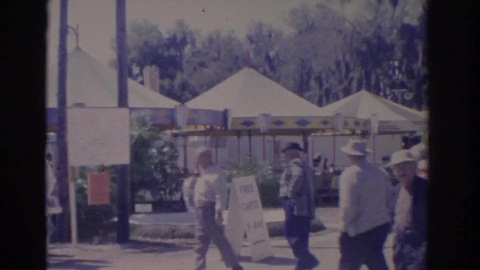 Florida carnival visitors in 1962 walk near a tented booth, a freestanding sign, and shaded trees at the edge of the fairground.
