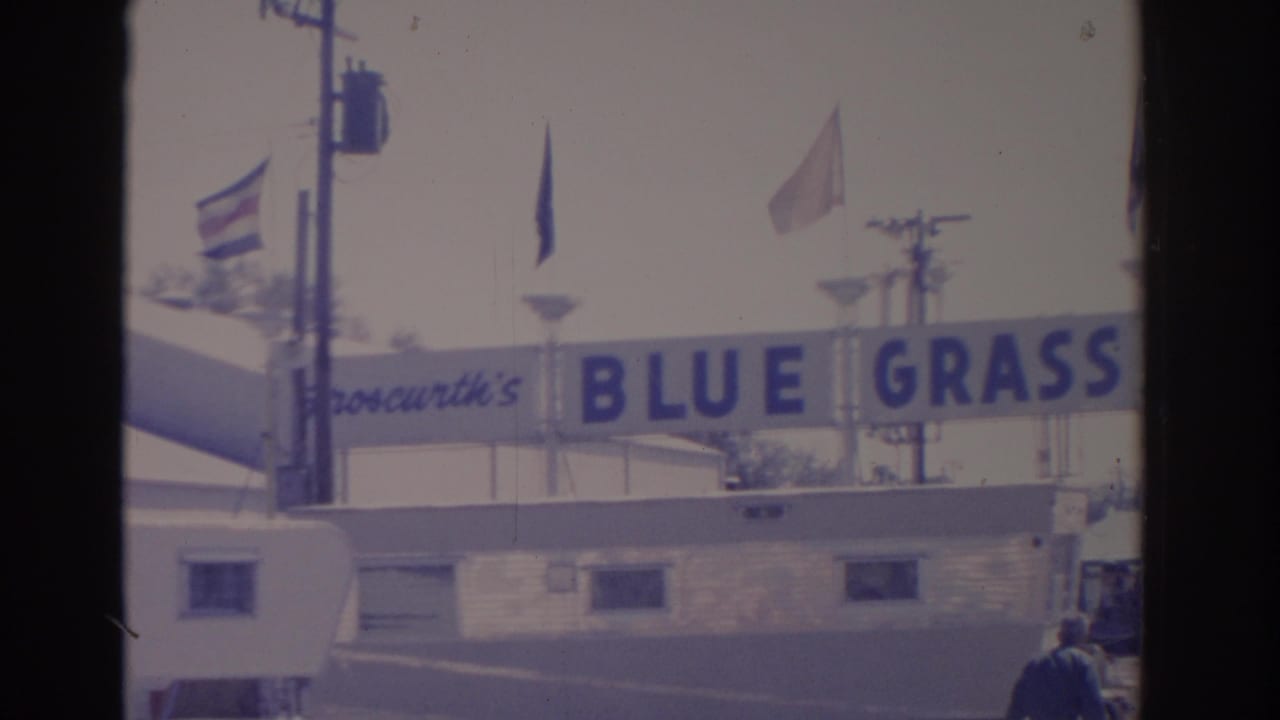 1962 Florida 8mm frame showing the Blue Grass Shows banner, flags, lampposts, and a white trailer at a fairground entrance.