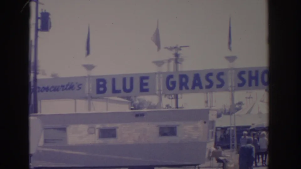 1962 Florida fairground view with the Blue Grass Shows sign over a trailer and a small group of visitors near the entrance.