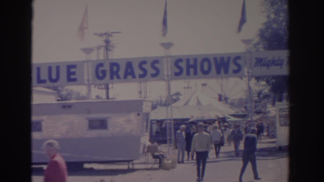 1962 Florida fairground shot with visitors moving past a trailer beneath the Blue Grass Shows banner and flags.