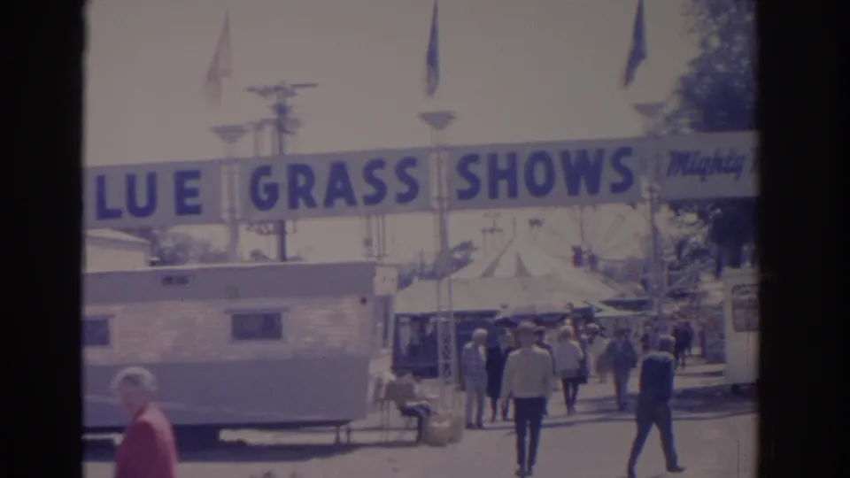 1962 Florida fairground shot with visitors moving past a trailer beneath the Blue Grass Shows banner and flags.