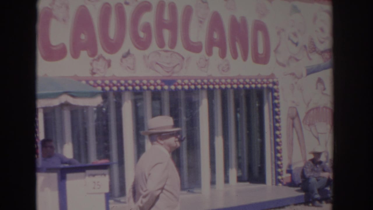 The suited man continues past the Laughland attraction, with painted carnival artwork and seated visitors visible nearby.