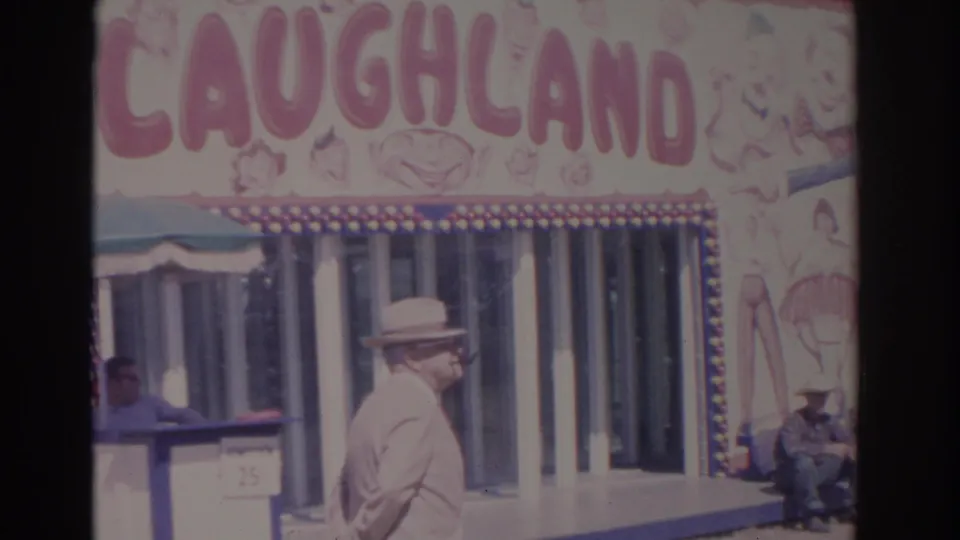 The suited man continues past the Laughland attraction, with painted carnival artwork and seated visitors visible nearby.