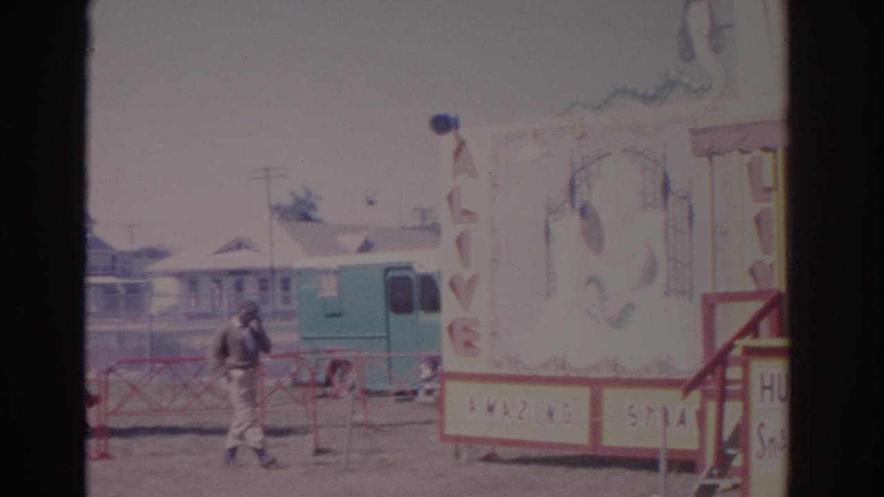 1962 Florida 8mm frame with a carnival showfront, a visible man, green trailer, red railing, and painted attraction panel.
