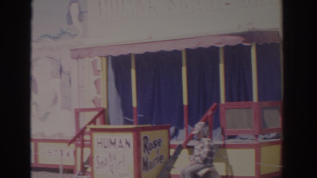 1962 Florida 8mm frame with the carnival booth entrance, Human Snake Girl and Rose Marie signs, blue curtains, and a seated visitor.