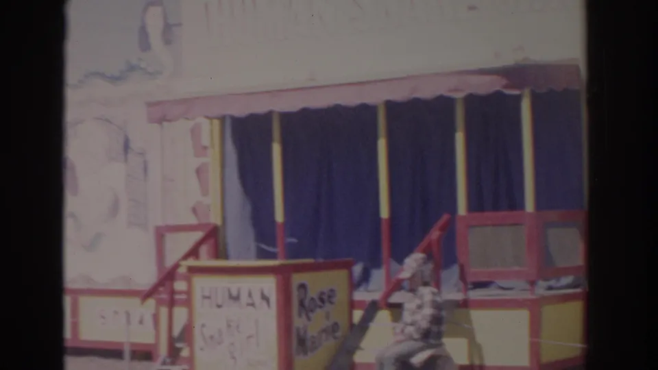 1962 Florida 8mm frame with the carnival booth entrance, Human Snake Girl and Rose Marie signs, blue curtains, and a seated visitor.