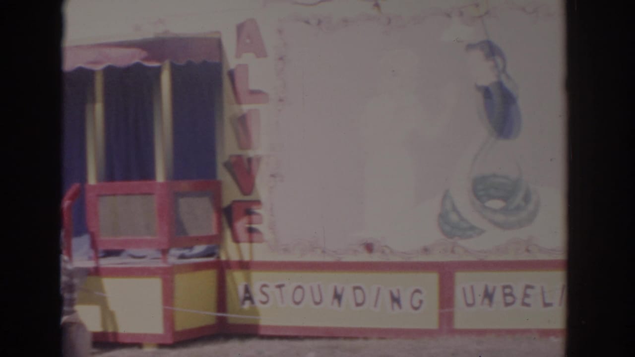 1962 Florida carnival booth facade with red trim, yellow panels, blue curtains, partial Astounding lettering, and vertical ALIVE sign.