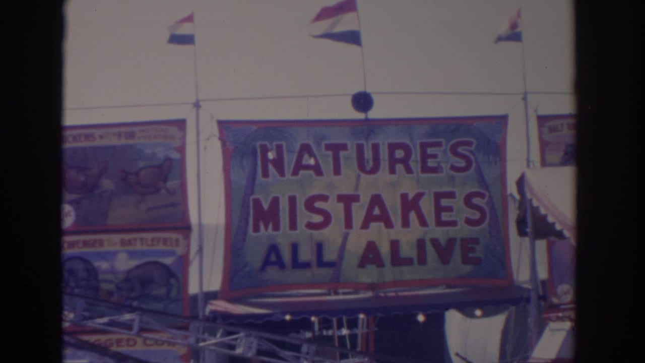 1962 Florida 8mm frame showing a large Natures Mistakes All Alive banner, surrounding painted sideshow panels, flags, and booth structure.