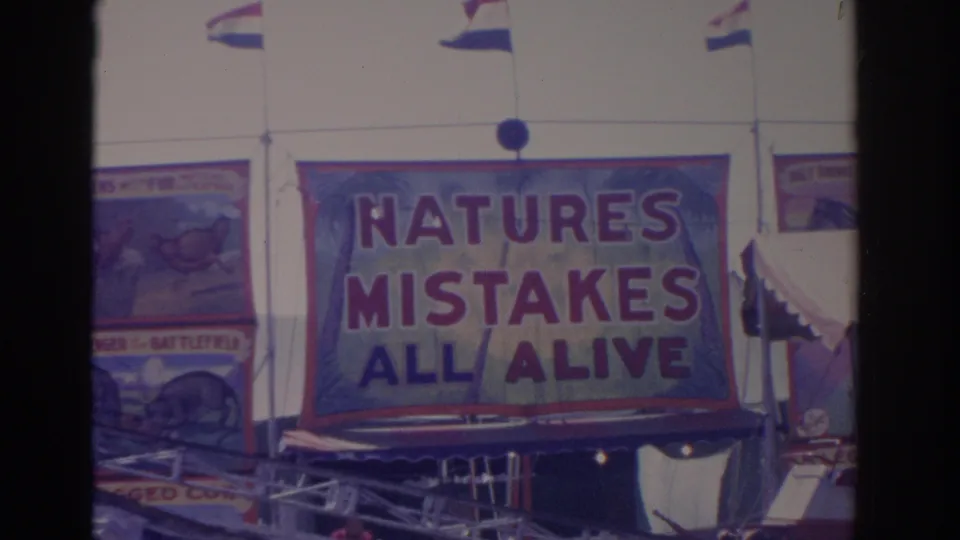 1962 Florida 8mm frame showing a large red and blue carnival banner reading NATURES MISTAKES ALL ALIVE with flags above.