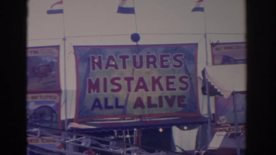 The home-movie frame centers on the NATURES MISTAKES ALL ALIVE sideshow banner, with flags and adjacent painted signs visible.