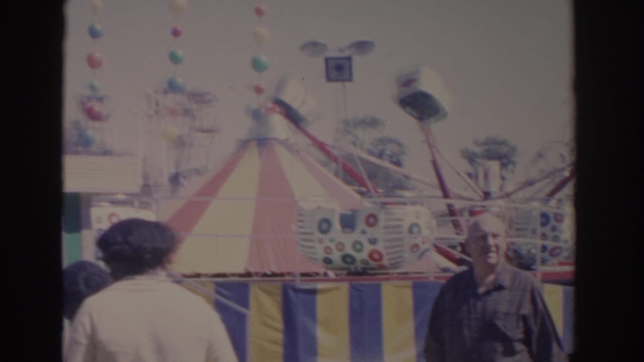1962 Florida midway view with adults walking past a striped tent and colorful carnival ride cars.