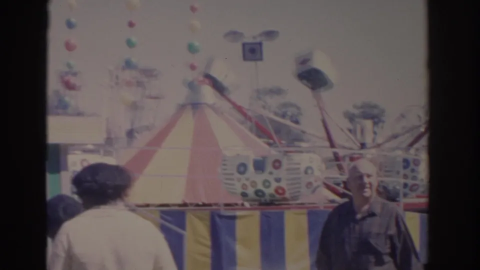 1962 Florida midway view with adults walking past a striped tent and colorful carnival ride cars.