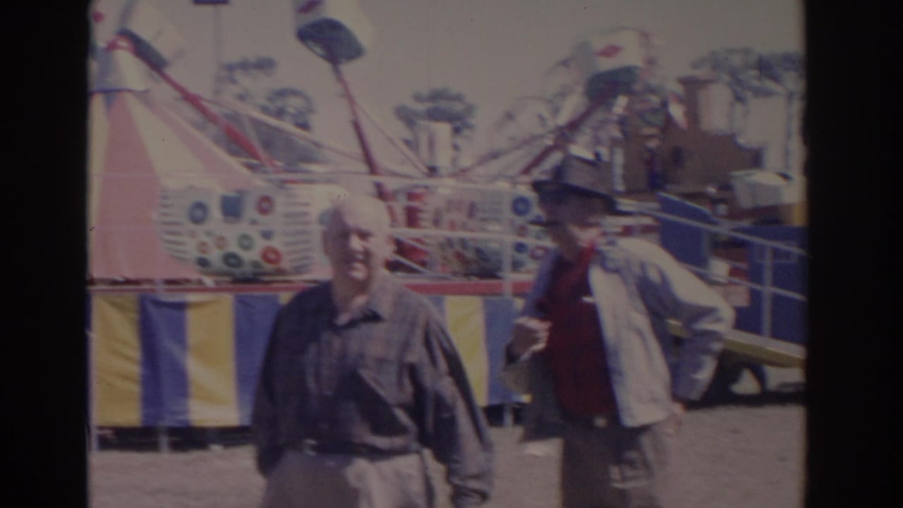 1962 Florida carnival frame with two adult men walking before a colorful ride and striped midway barriers.