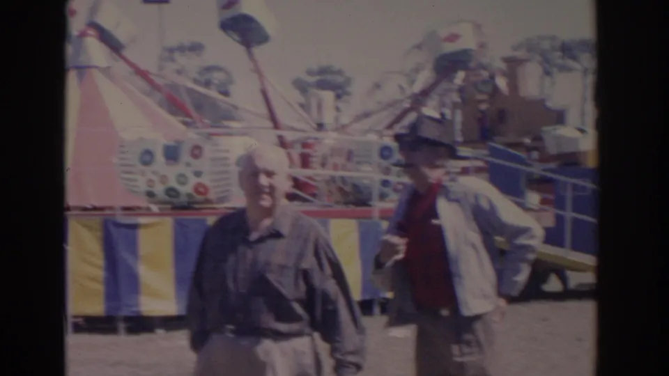 1962 Florida carnival frame with two adult men walking before a colorful ride and striped midway barriers.