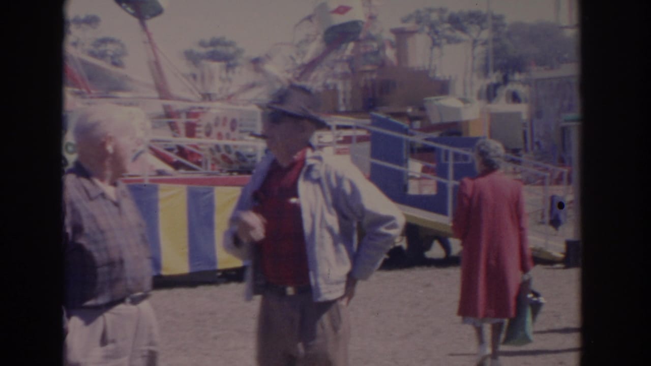 1962 Florida home-movie frame showing adults in hats and dresses walking past striped barriers and fairground rides.