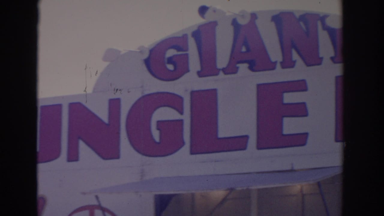 1962 Florida 8mm frame showing a cropped Giant Jungle sign across a pale attraction facade with bulb details and a lower entrance area.
