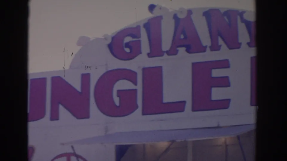 1962 Florida 8mm frame showing a cropped Giant Jungle sign across a pale attraction facade with bulb details and a lower entrance area.