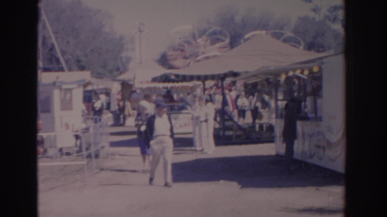 Visitors walk past a sunlit amusement park stand with awnings and decorations.