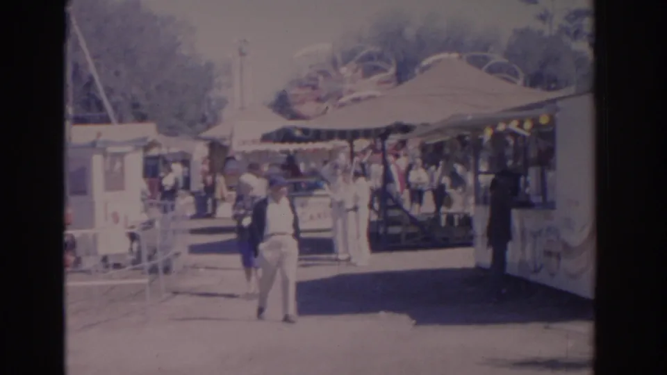 1962 Florida home-movie frame showing visitors on a bright amusement park walkway near a shaded midway stand.