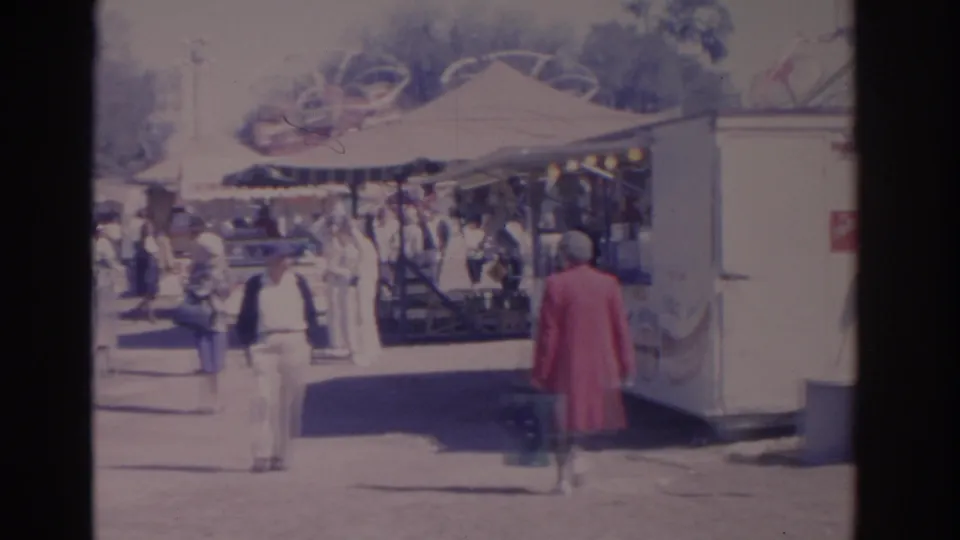1962 Florida park scene with visitors moving past a shaded concession or game stand and a person in a red coat at the edge.