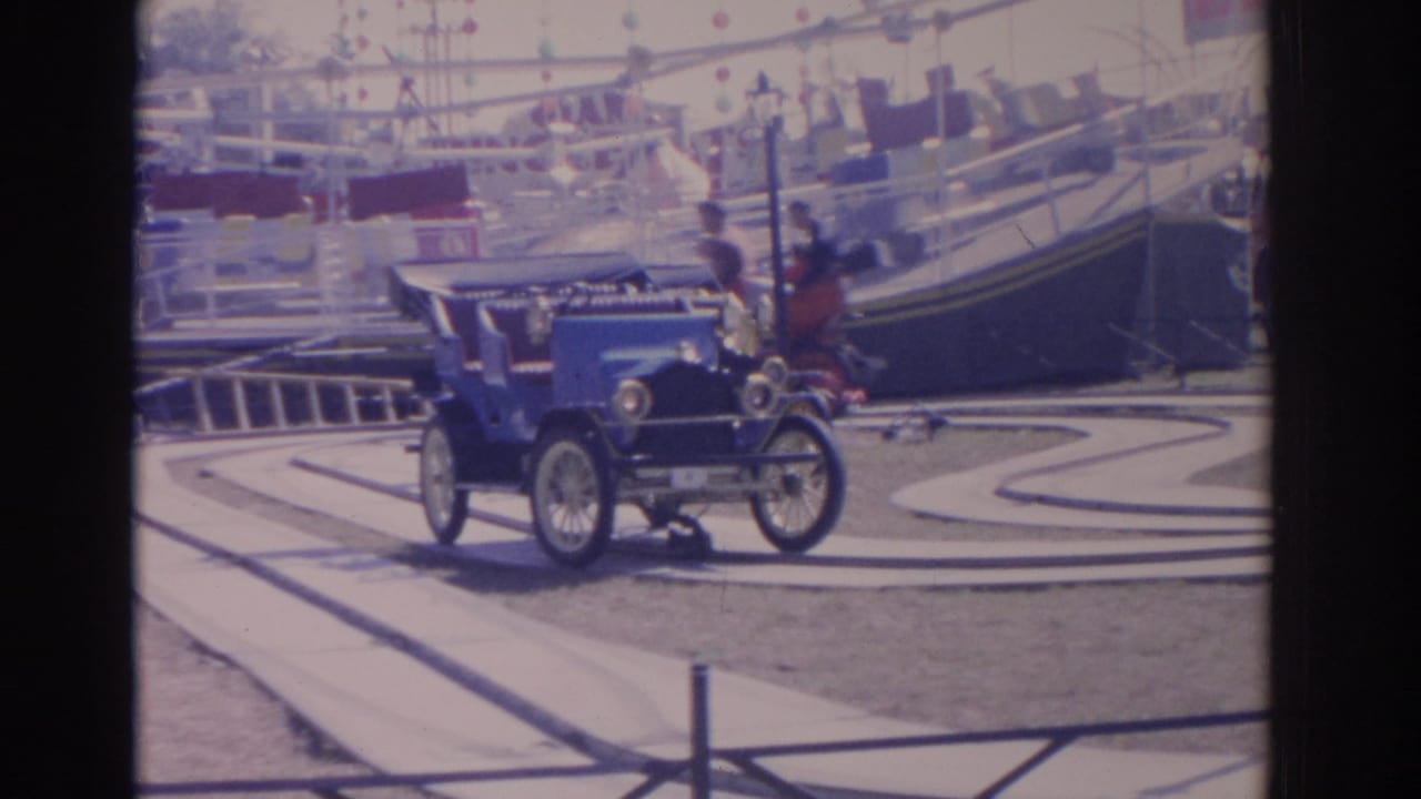1962 Florida 8mm frame showing a blue antique-style amusement ride car on a curving track with railings and ride structures in the background.