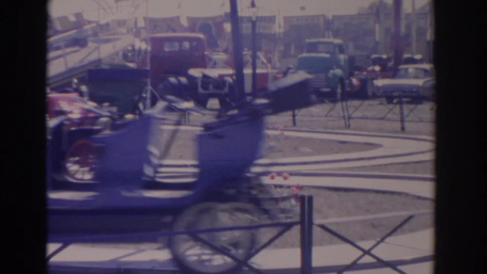 A blue antique car blurs past the foreground during a 1962 Florida outdoor automobile display, with parked vehicles and tents behind it.