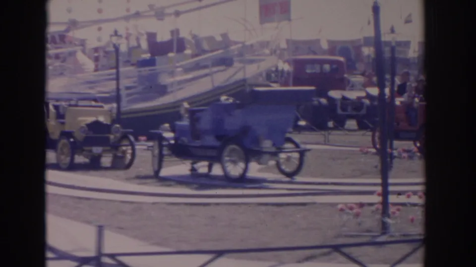 The blue antique car continues across the circular display area, with other early automobiles, railings, and fair tents in the background.