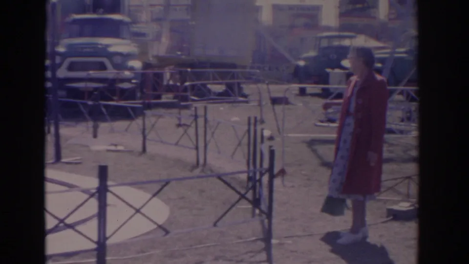 1962 Florida home-movie frame showing a woman in a red coat near metal barriers, with parked vehicles and fairground activity in the background.