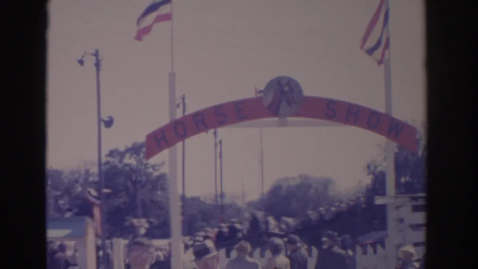 1962 Florida frame with spectators gathered under a red HORSE SHOW entrance arch, American flags, trees, and white fencing nearby.