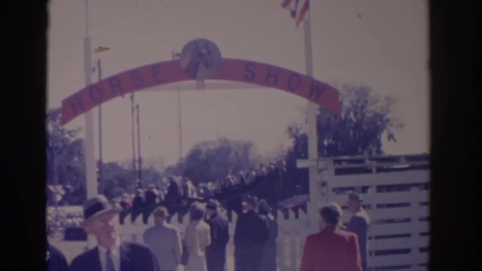 1962 Florida frame showing people in mid-century clothing near the HORSE SHOW arch, with white fencing, flags, and trees at the entrance.