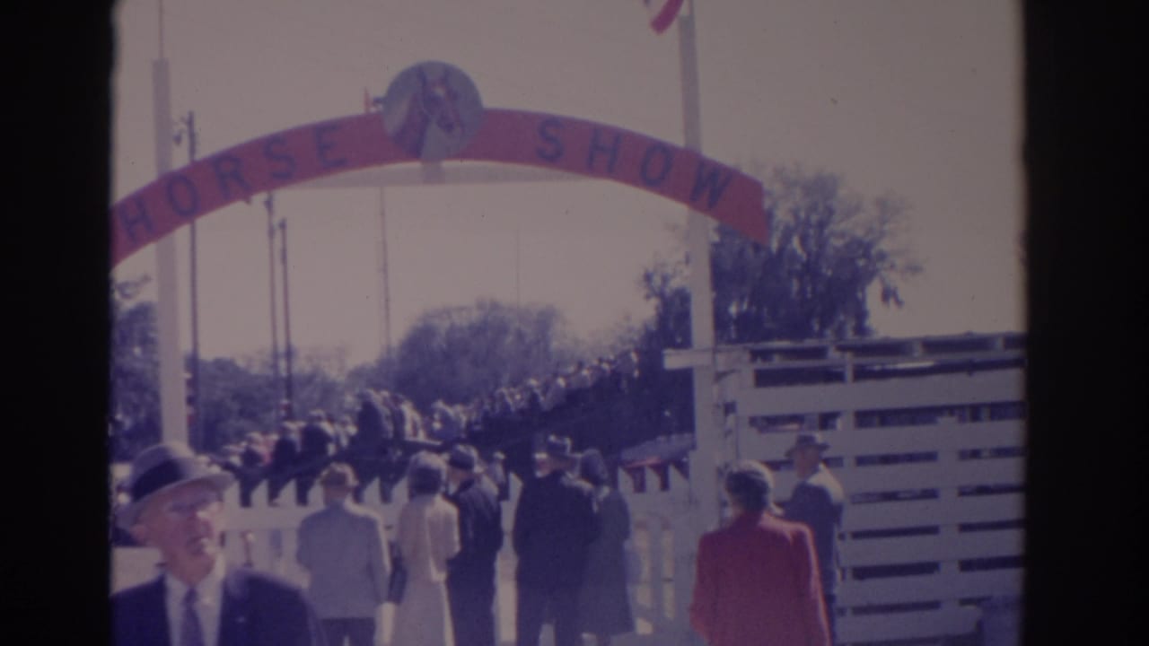 Crowd walking under a red HORSE SHOW arch beside a white fence.