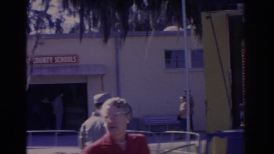 A woman in red stands near the camera as fairgoers pass a building marked SCHOOLS at a Florida fair in 1962.