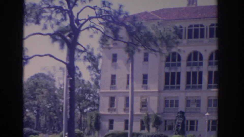 1962 Florida home-movie view of a multi-story school building with arched windows, trees, poles, and bright daylight exposure.