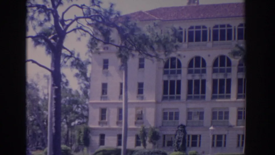 1962 Florida school exterior with tall arched windows, tree branches, a pole, and soft 8mm color rendering.