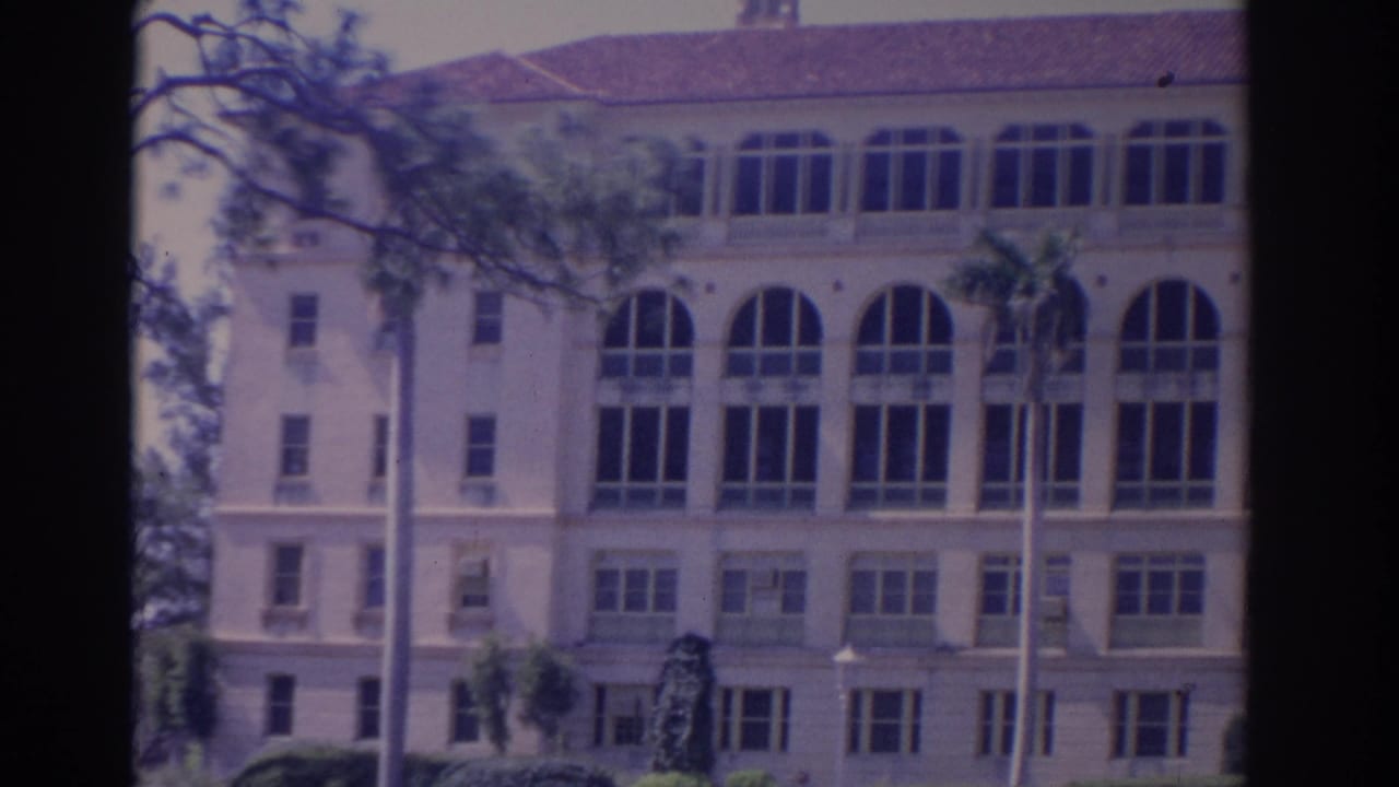 1962 Florida 8mm frame showing a large cream building with arched windows, a red tile roof, palms, and landscaped foreground.