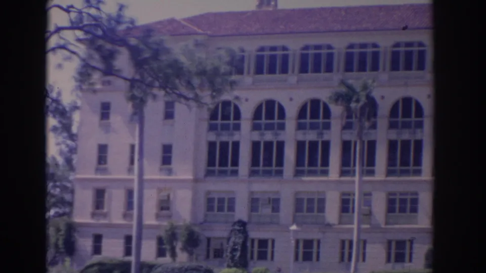 1962 Florida 8mm frame showing a large cream building with arched windows, a red tile roof, palms, and landscaped foreground.