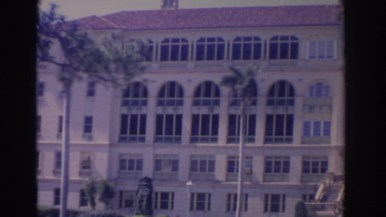Large building facade with rows of arched windows, shutters, palms, and red tile roof.